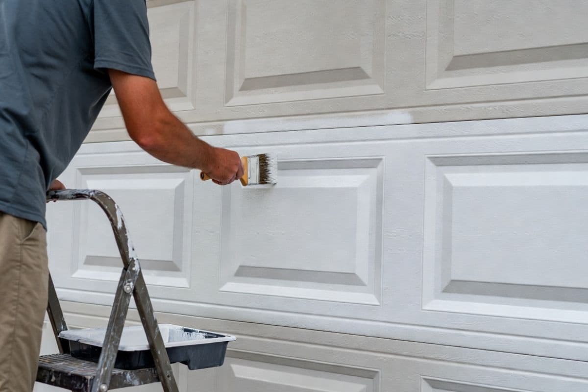 Homeowner painting a garage door panel with a brush and paint tray on a step ladder
